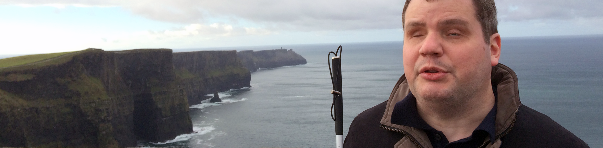 A blind man poses in front of a view of the ocean and cliffs.